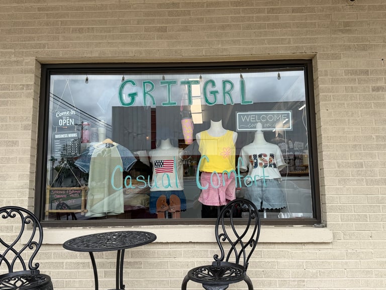 Storefront window display for GRITGRL boutique with mannequins wearing clothing on tan brick wall, decorative chairs below