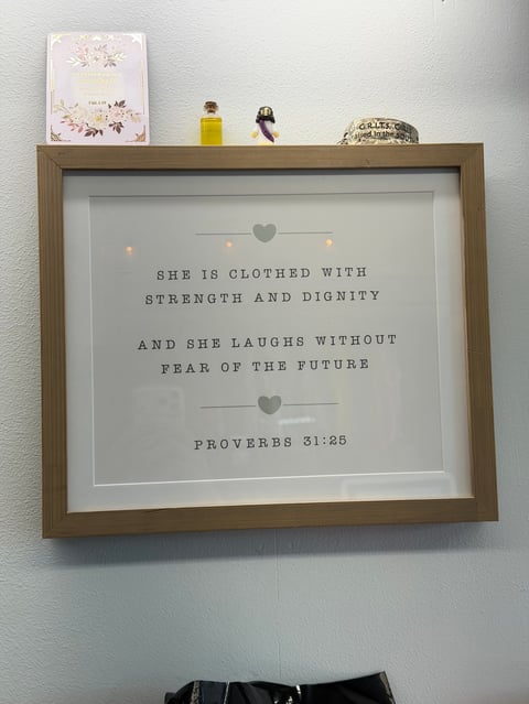 Framed inspirational quote from Proverbs 31:25 displayed on white wall with decorative items on shelf above