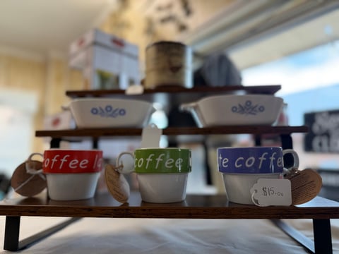 Three white coffee cups with colored bands labeled coffee in red, green, and blue, displayed on a shelf with a coffee maker in the background